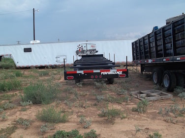Two flatbed trailers parked on a dirt lot with a building in the background.
